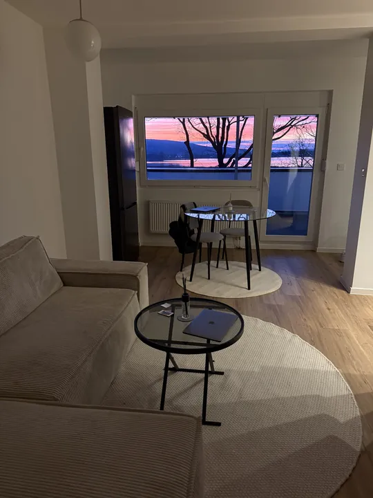 Wohnraum mit Sofa, rundem Glastisch und Fensterfront zum See im Abendrot